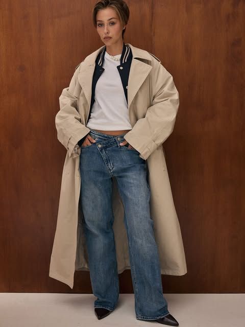 Model in beigem Trenchcoat und weiter Jeans vor einer Holzwand